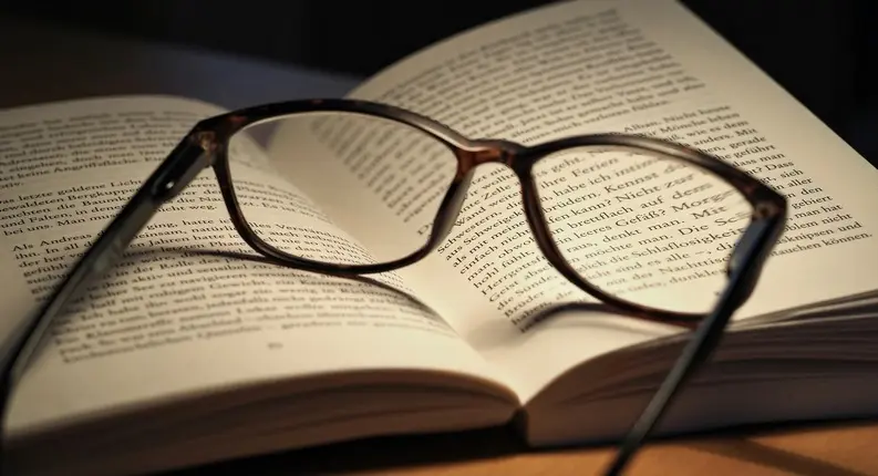 Tortoiseshell eyeglasses resting on an open book on a wooden table, lit by warm side light